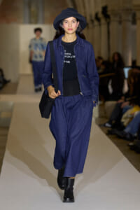 Model walks the runway in a dark blue denim suit with a matching wide-brim hat and black top, carrying a black shoulder bag.