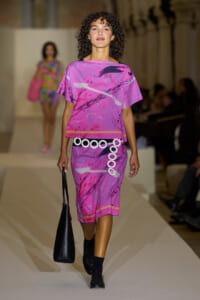 Model walks a runway in a pink-purple abstract print dress with a white circular belt, carrying a black bag on her left side.