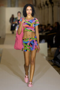 Model walks the runway in a neon multicolor geometric crop top and matching shorts, with a pink tote on her shoulder.