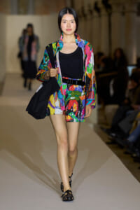 Model walking a fashion runway in a colorful abstract-print jacket and matching shorts, black top, belt, and sandals, carrying a black tote bag.