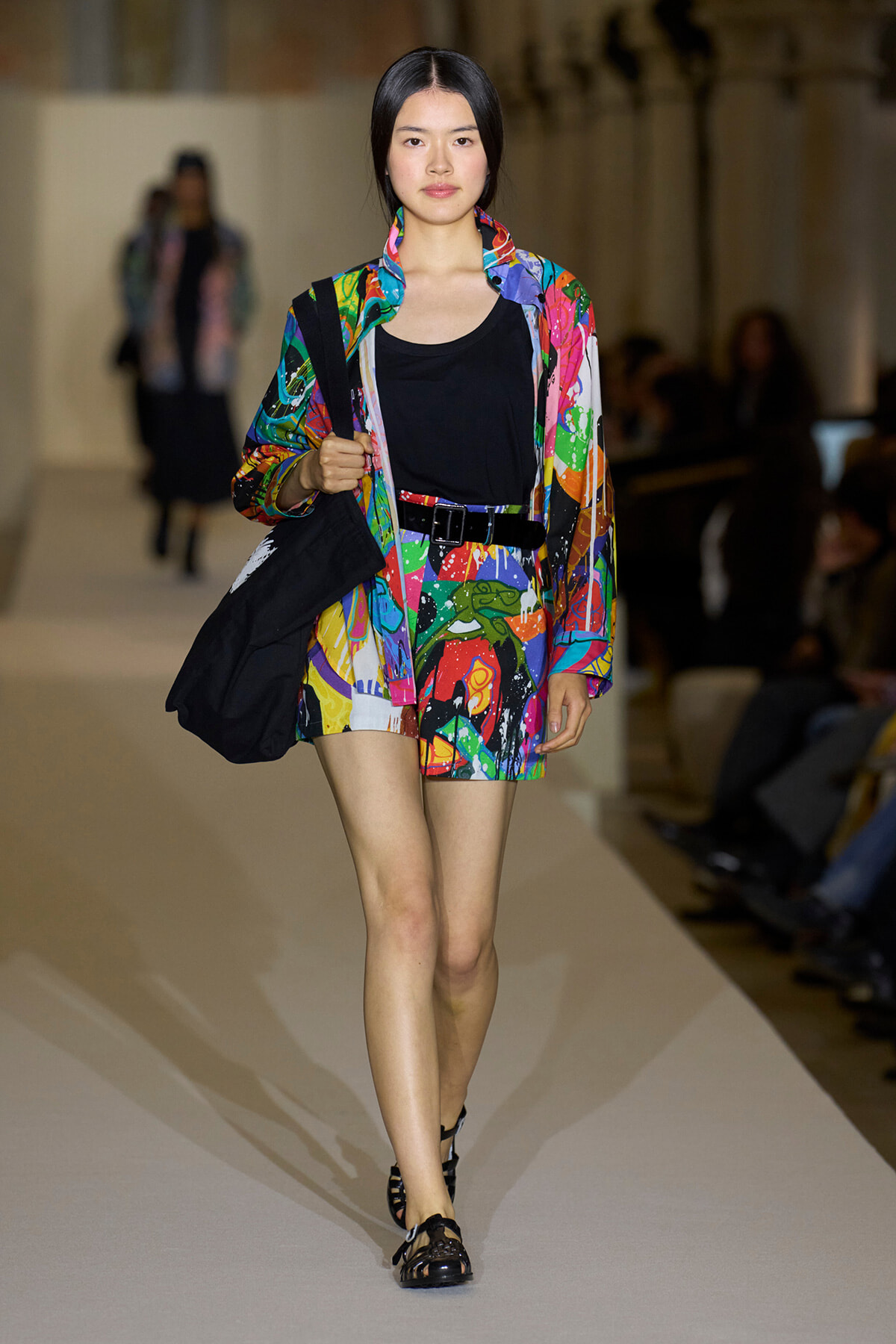 Model walking a fashion runway in a colorful abstract-print jacket and matching shorts, black top, belt, and sandals, carrying a black tote bag.