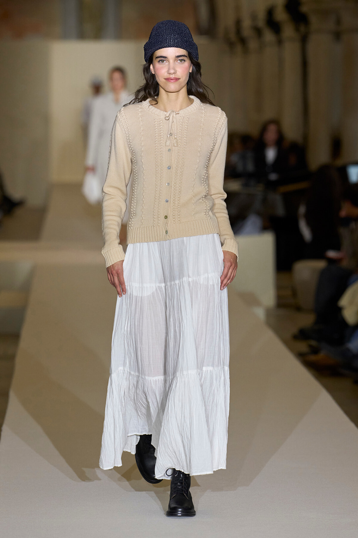 Model on a runway wearing a beige knit cardigan, white tiered maxi skirt, black lace-up boots, and a navy beanie stitch cap.