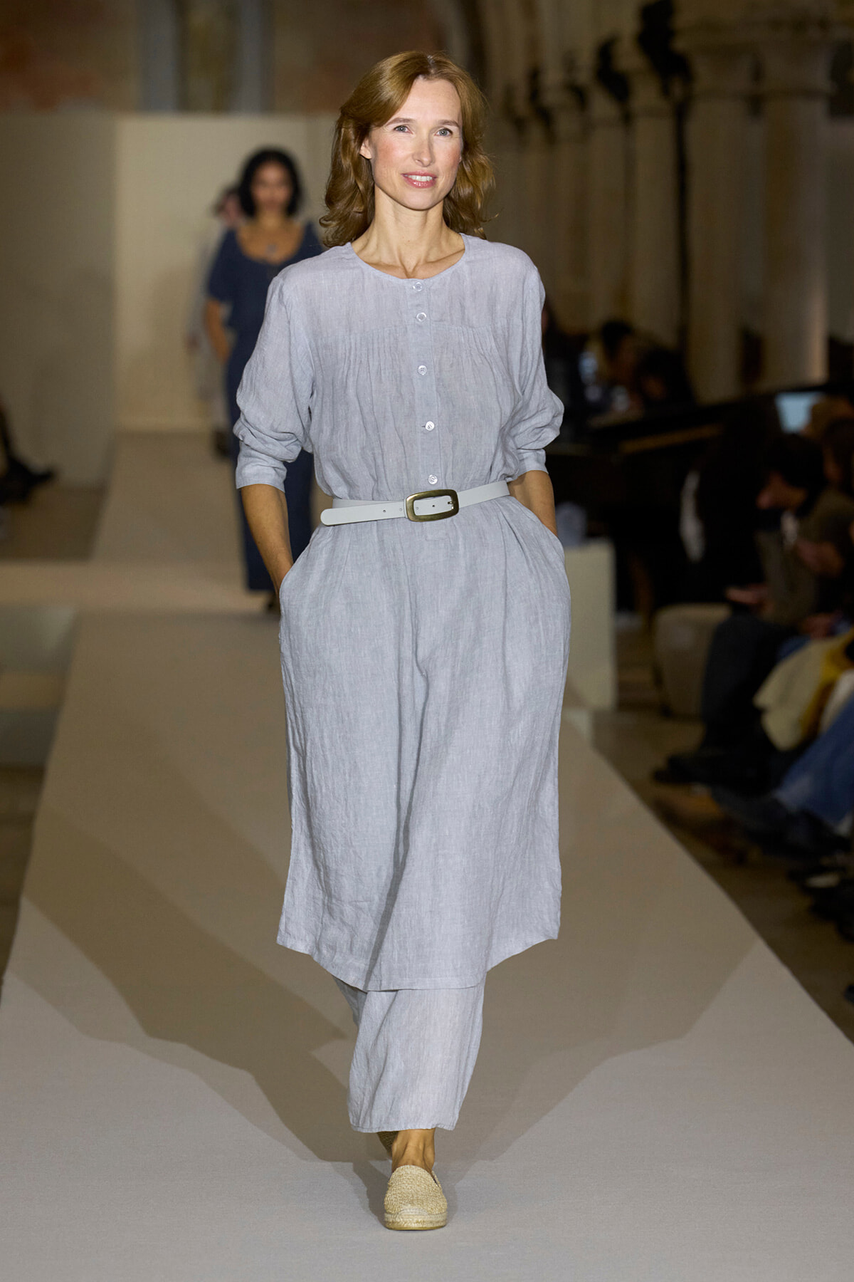 Woman models a light gray linen shirt-dress belted at the waist, matching wide-leg pants, walking a runway in espadrilles.