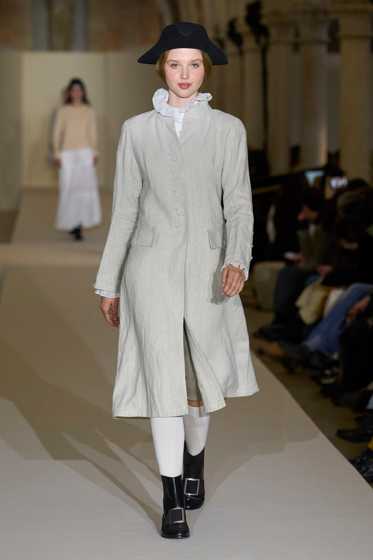 Model walks a fashion runway wearing a light grey knee-length coat with large buttons, white ruffled blouse, black wide-brim hat and buckled boots in a minimalist show setting.