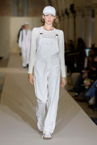 Model walks the runway in white utility overalls, cap and sneakers on a pale beige backdrop.