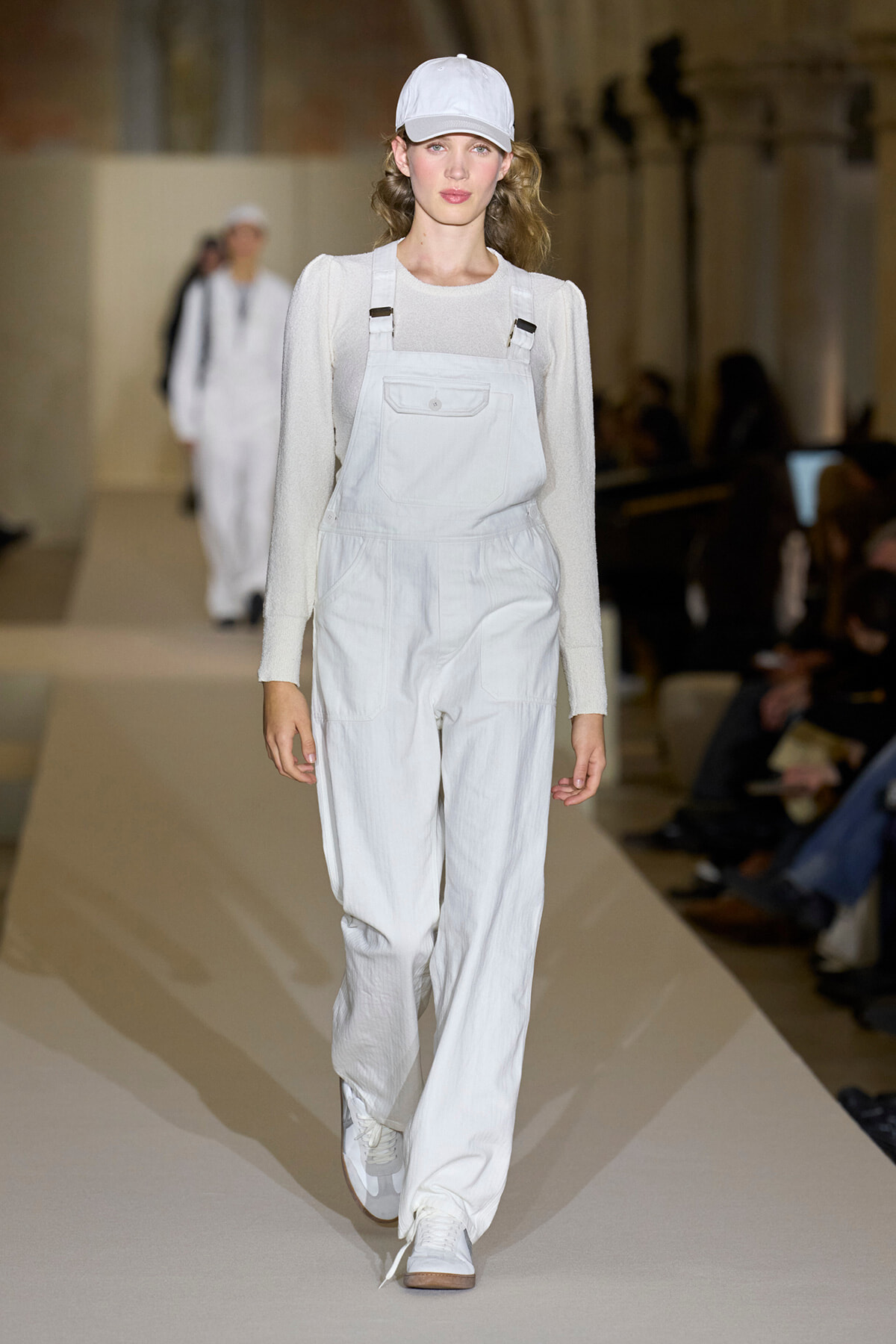 Model walks the runway in white utility overalls, cap and sneakers on a pale beige backdrop.