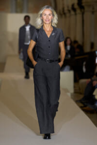Model walks a fashion show runway in a charcoal gray short-sleeve jumpsuit with a black belt and shoulder bag, hands in pockets.