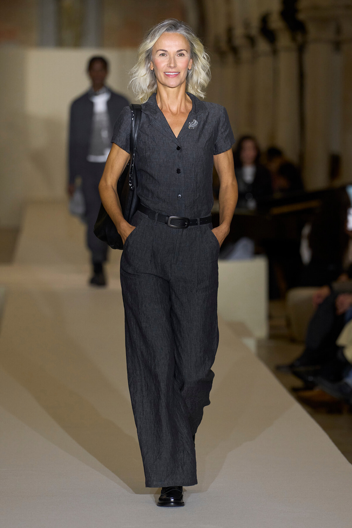 Model walks a fashion show runway in a charcoal gray short-sleeve jumpsuit with a black belt and shoulder bag, hands in pockets.