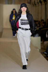 Model walking a runway in a white cargo jumpsuit look with a black bomber jacket, graphic tee, and black cap.