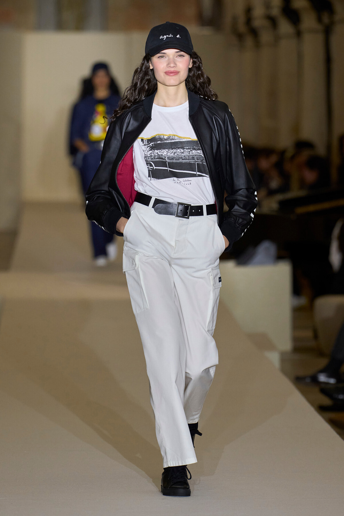 Model walking a runway in a white cargo jumpsuit look with a black bomber jacket, graphic tee, and black cap.