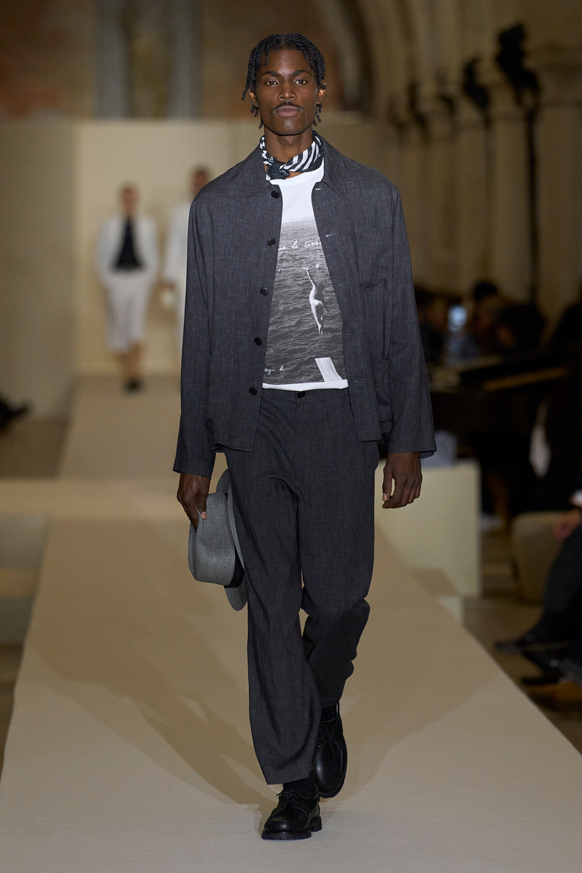Male model walks a runway in a dark gray tailored jacket and matching wide-leg trousers, layered over a graphic white T-shirt, with a striped scarf and black lace-up boots, carrying a gray hat.
