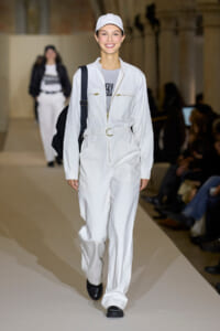 Female model walks runway in a white utility jumpsuit with belt, cap, and black backpack.