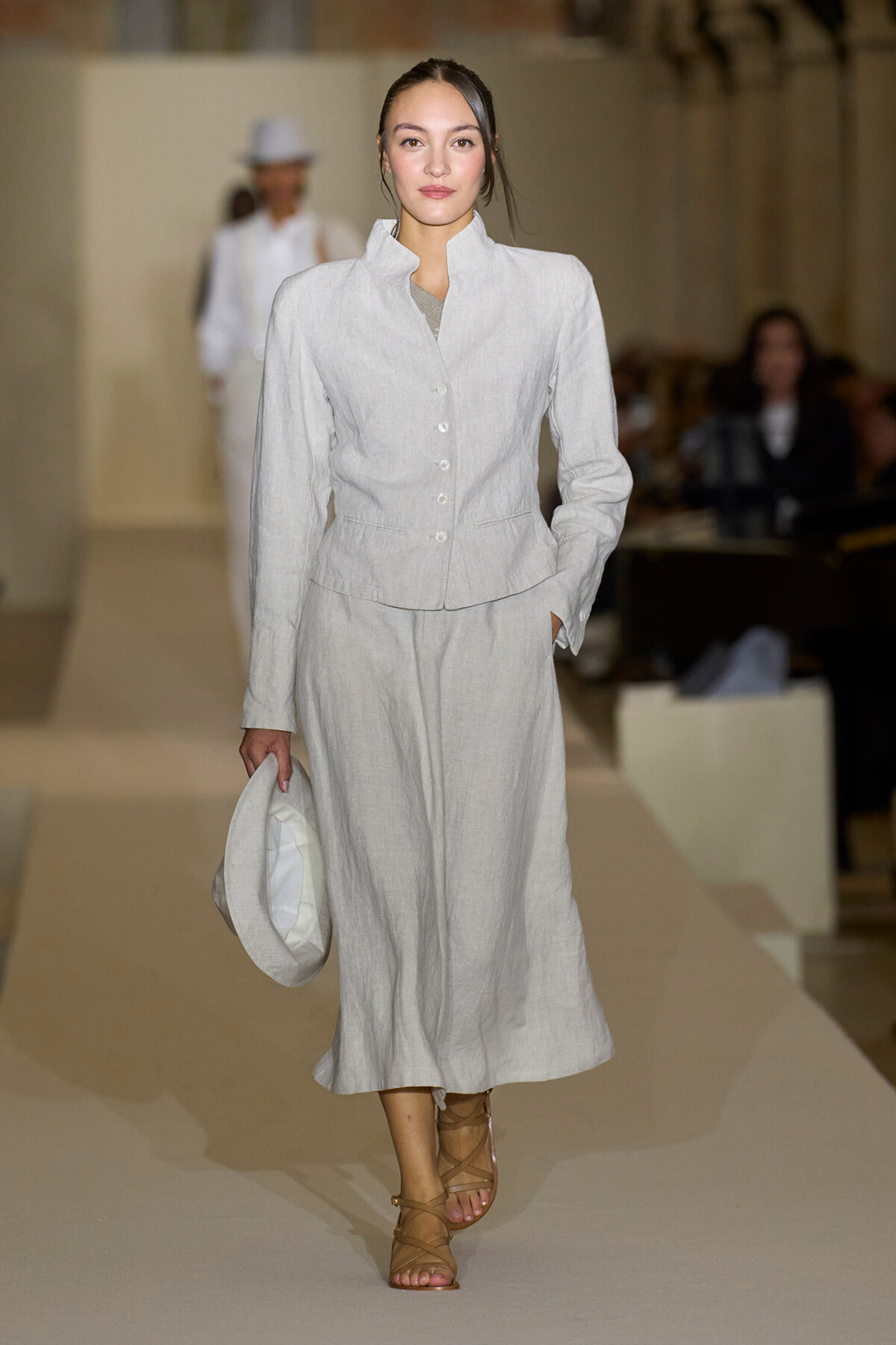 Model on a runway wearing a pale gray linen suit with buttoned jacket and long skirt, holding a white hat at her side.