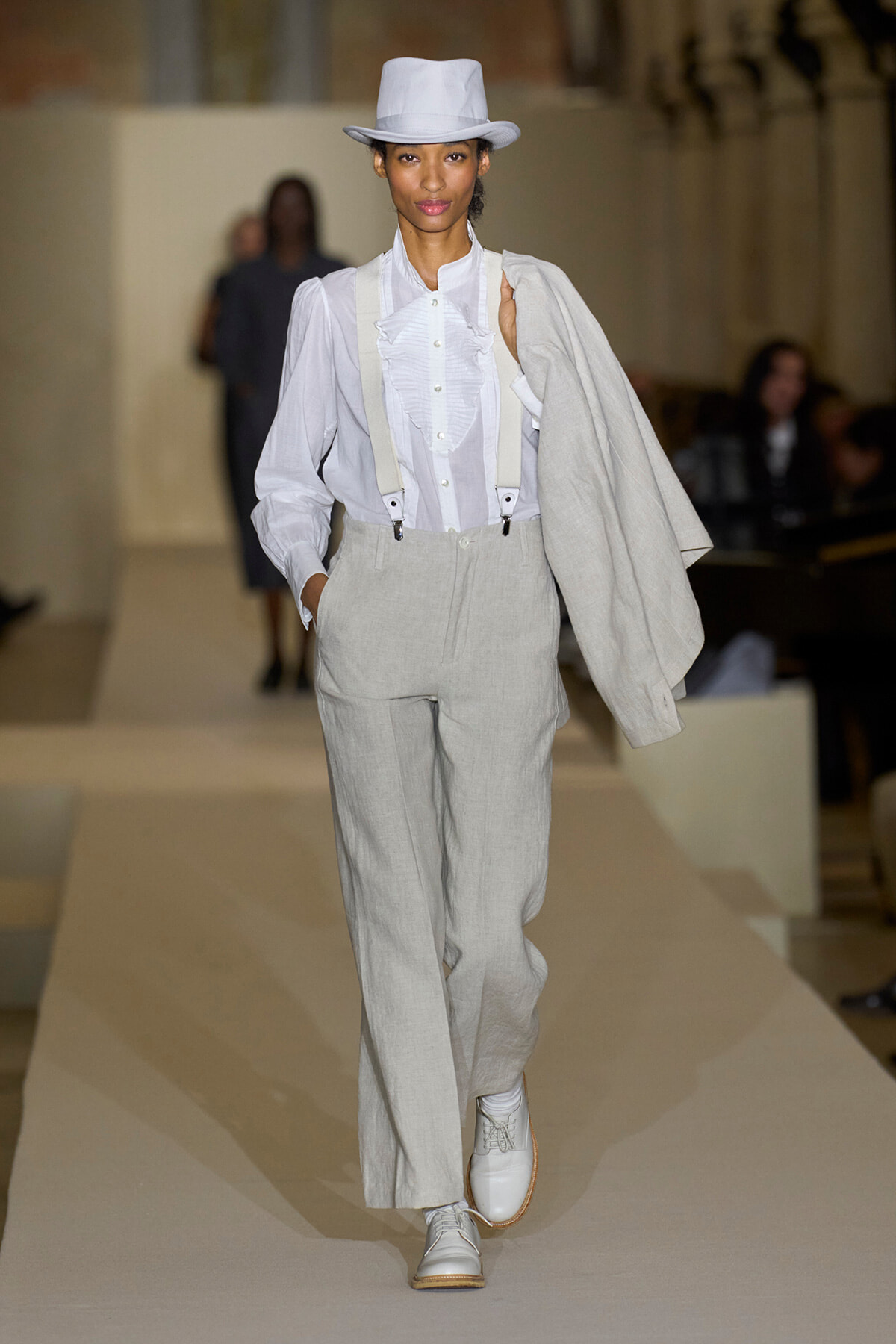 Model walks a runway in a light gray suit with suspenders, white ruffled shirt, white hat, and matching shoes; coat draped over her shoulder.
