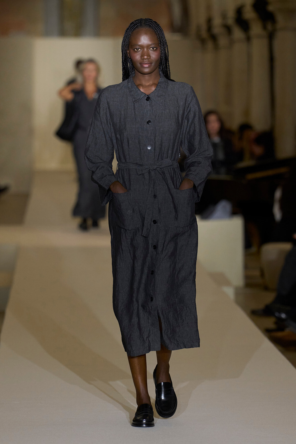 Model walking a fashion runway wearing a knee‑length dark gray belted shirt dress with pockets and black loafers.