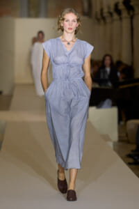 Model walking the runway in a light blue-gray jumpsuit with a cinched waist and pockets, paired with burgundy flats and a pendant necklace.