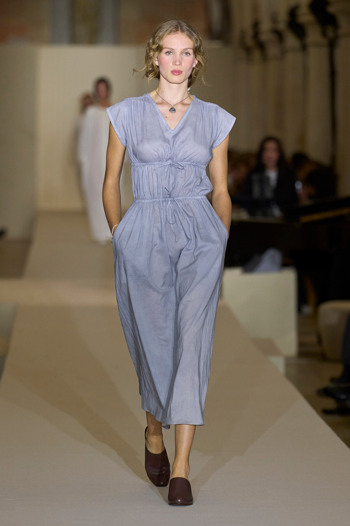 Model walking the runway in a light blue-gray jumpsuit with a cinched waist and pockets, paired with burgundy flats and a pendant necklace.