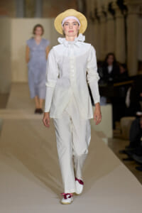 Model on a runway wearing a cream white ensemble: a shirt with a large pleated ruffle collar, matching wide-leg trousers, and a straw hat.