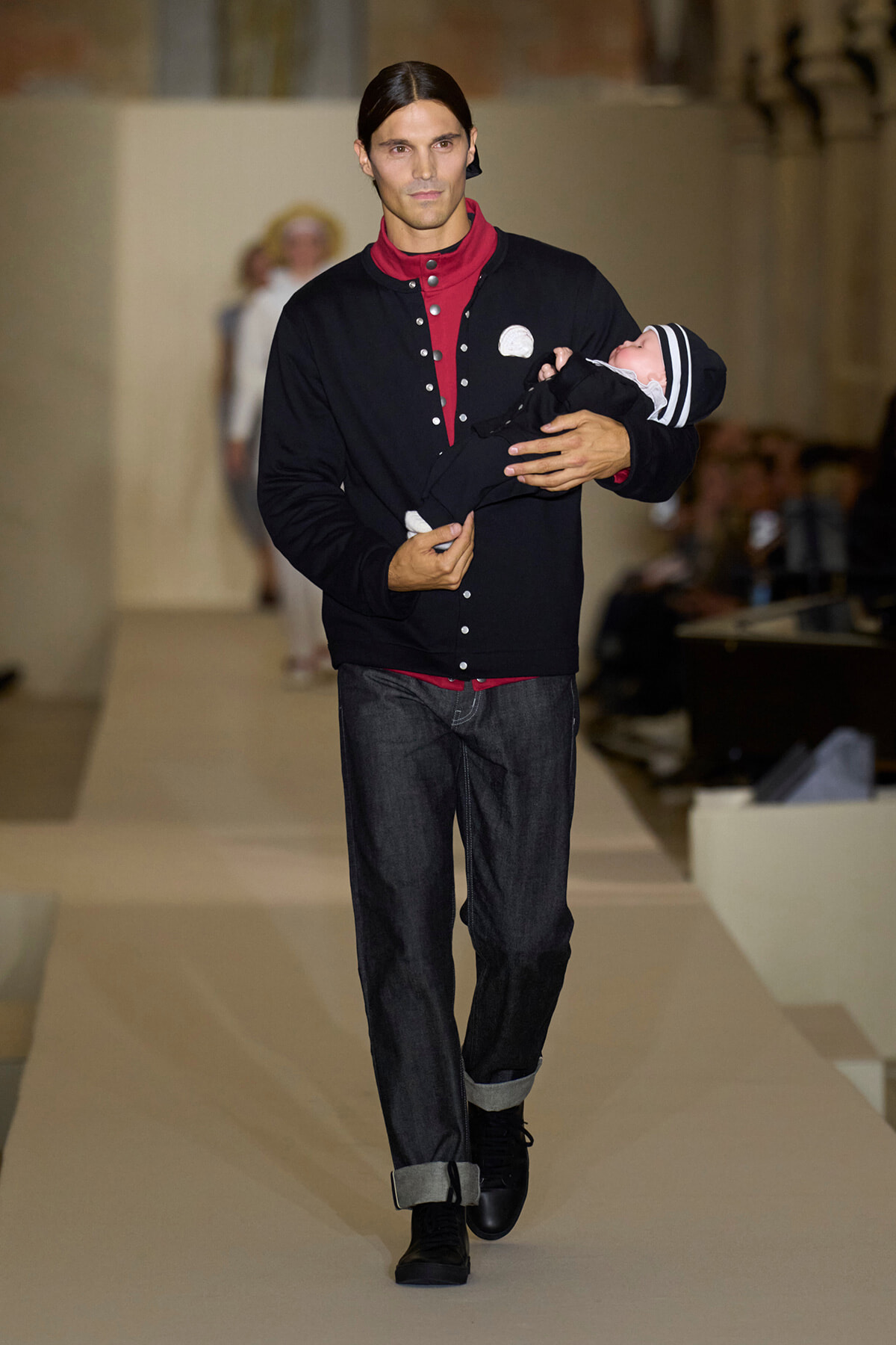 Male model in a black cardigan and red shirt walks a runway while cradling a baby dressed in black with a striped hat, audience visible in background.