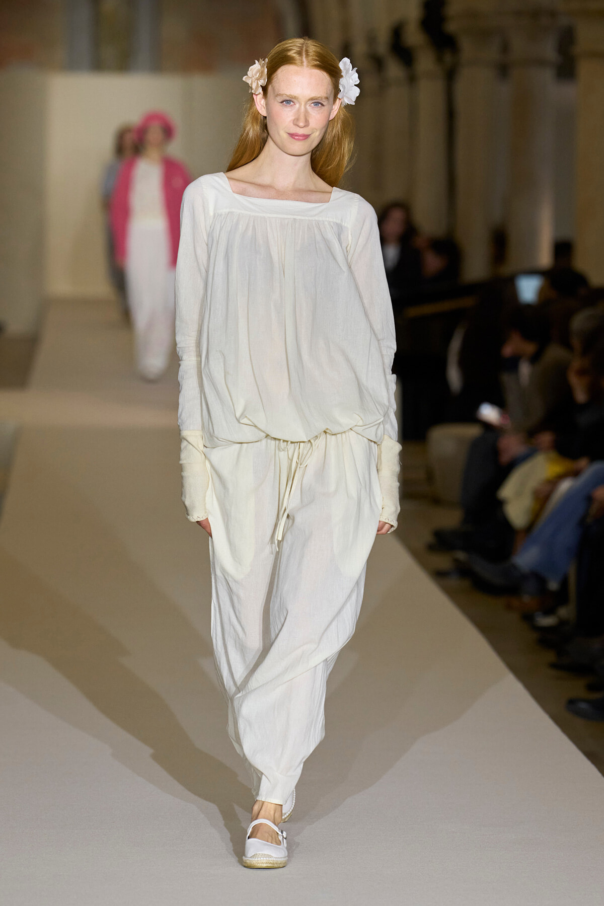 Model walks the runway in an oversized white outfit with a drawstring waist, white sandals, and white flowers in her hair.