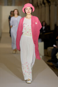Model walking a runway in a pink cardigan and hat over a white floral-print jumpsuit/dress with white shoes.