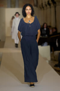 Model walks a fashion runway in a navy blue two-piece outfit with a ruffled off-the-shoulder top and wide-leg pants, wearing a shell pendant necklace.