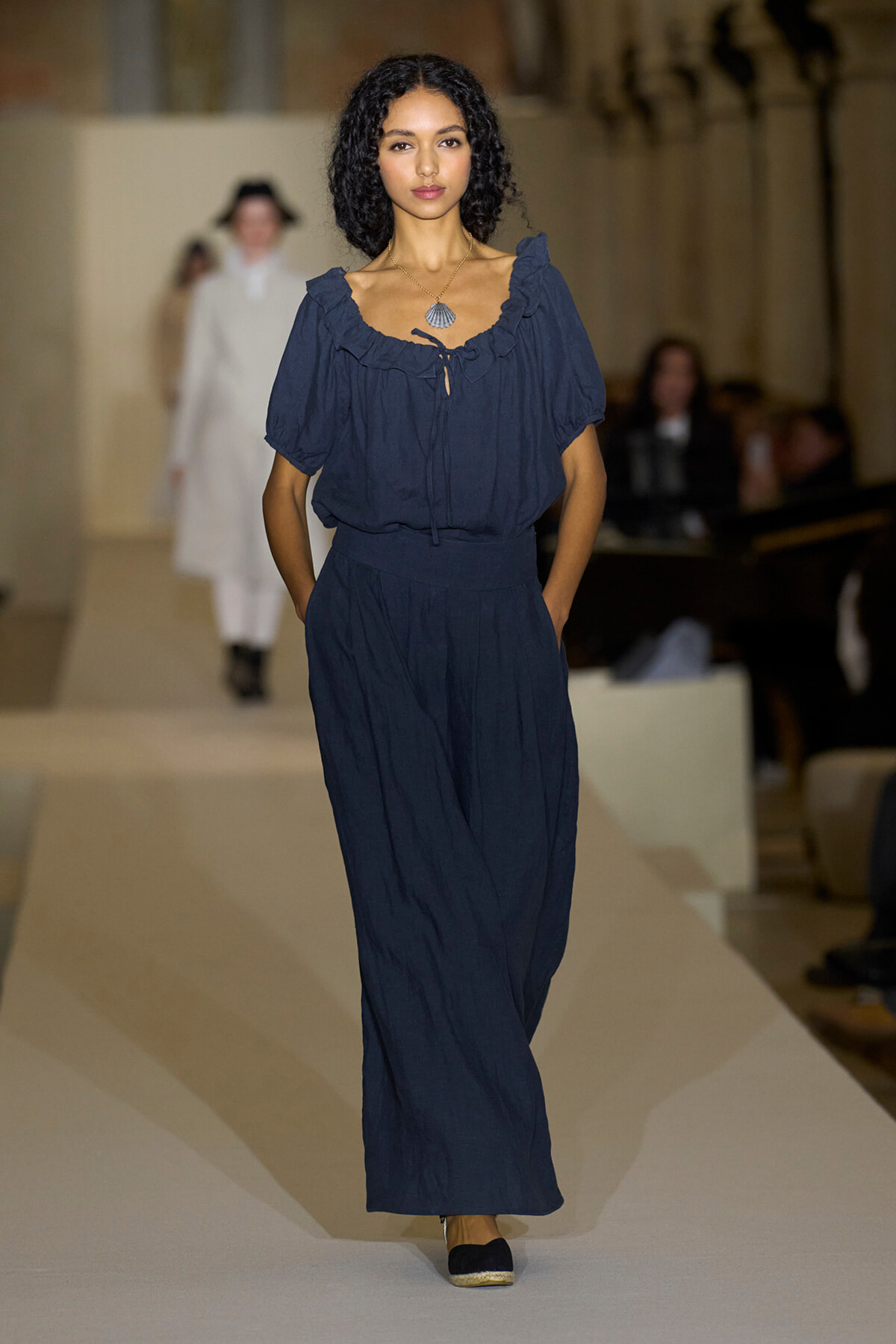 Model walks a fashion runway in a navy blue two-piece outfit with a ruffled off-the-shoulder top and wide-leg pants, wearing a shell pendant necklace.