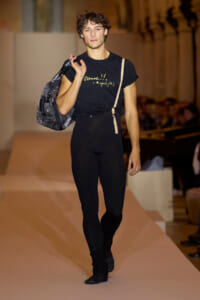 Male fashion model walking a runway while carrying a gray backpack over his shoulder; wearing a black t-shirt with yellow text and black leggings, beige strap visible, audience in background. Includes shirt text: 'Danse!! agnès b.'