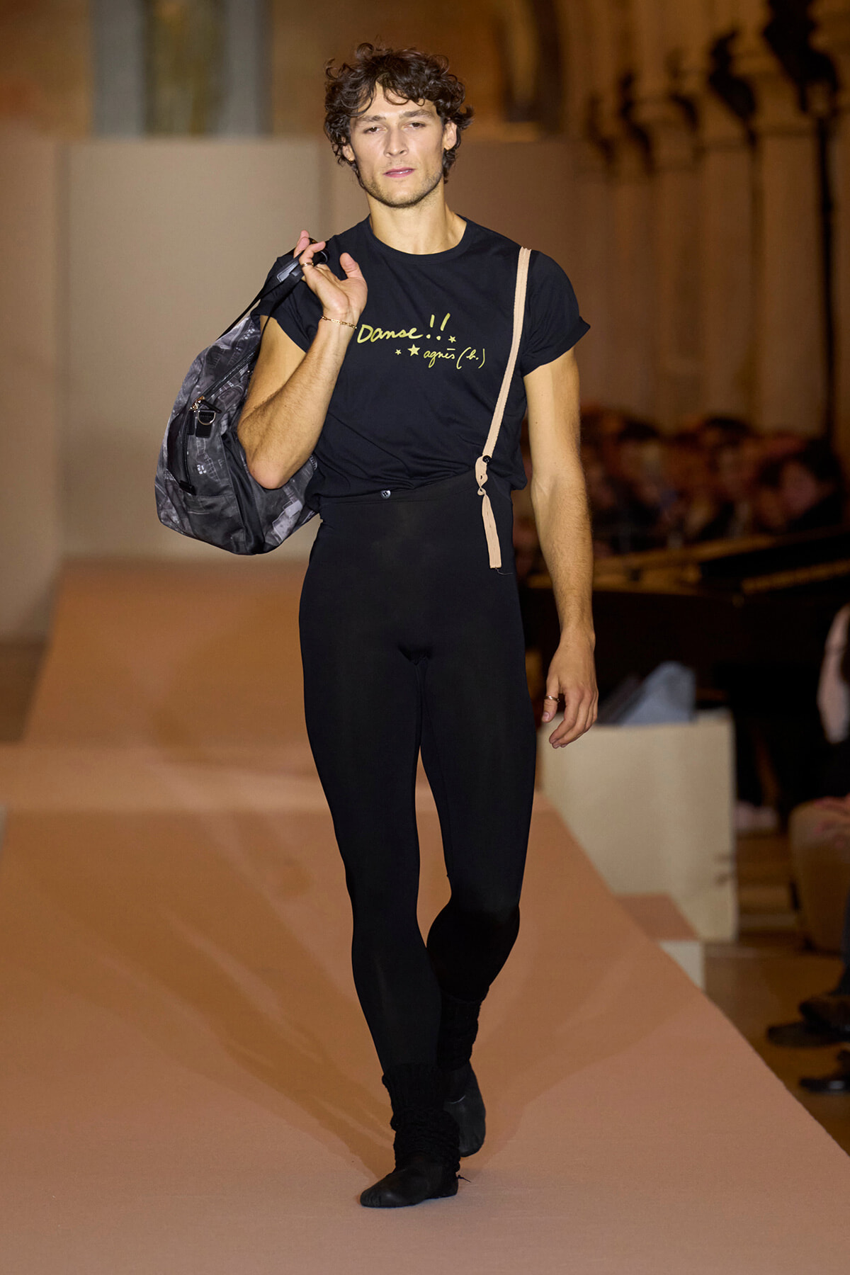 Male fashion model walking a runway while carrying a gray backpack over his shoulder; wearing a black t-shirt with yellow text and black leggings, beige strap visible, audience in background. Includes shirt text: 'Danse!! agnès b.'