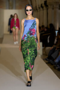 Model walks the runway in a sleeveless, gradient blue-to-green floral dress with pink flowers; black platform sandals and light sunglasses add a chic, summery look.