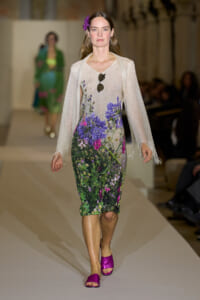 Model on a runway wearing a knee-length floral dress with purple and pink blossoms, a pale sheer cardigan, purple sandals, sunglasses hanging from the dress, and a purple flower in her hair.
