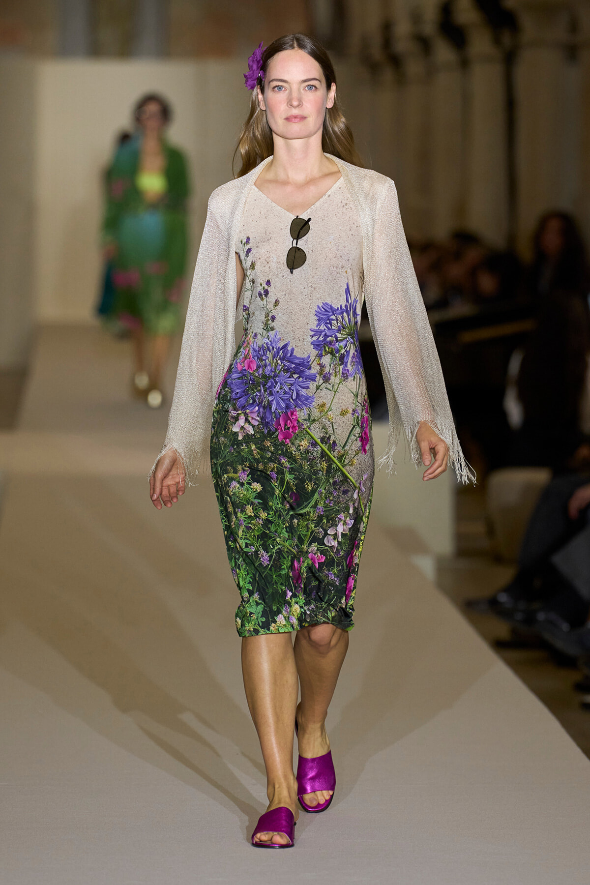 Model on a runway wearing a knee-length floral dress with purple and pink blossoms, a pale sheer cardigan, purple sandals, sunglasses hanging from the dress, and a purple flower in her hair.