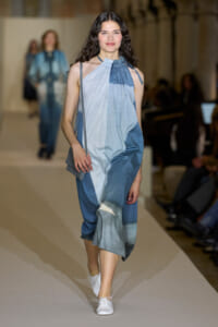 Runway model walking in a sleeveless blue gradient halter dress with a small gray purse and white sneakers.