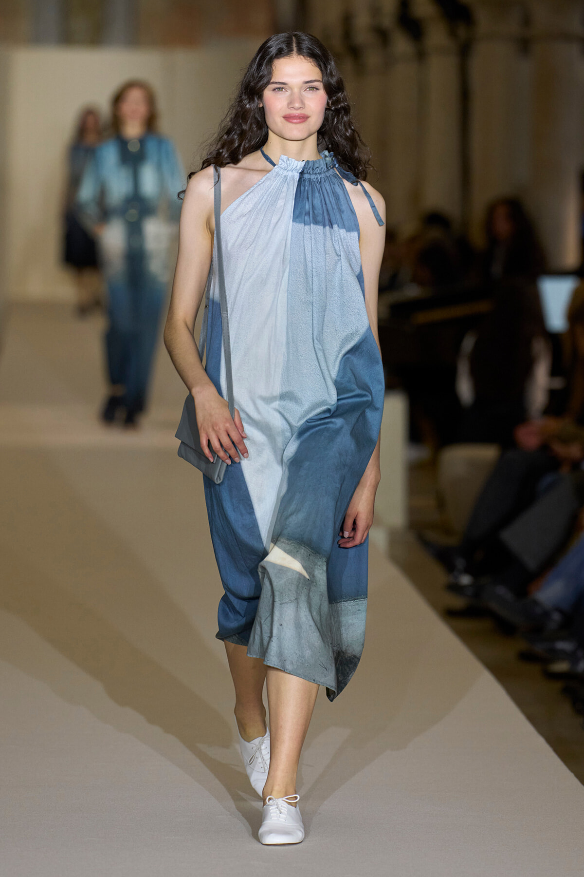 Runway model walking in a sleeveless blue gradient halter dress with a small gray purse and white sneakers.