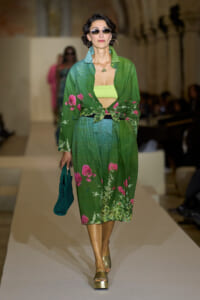 Model walks the runway in a green floral coordinated set with a tied waist and neon lime bandeau, gold platform sandals, and a teal bag, wearing sunglasses.