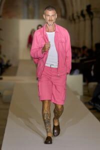 Male model walking a runway in a pink cargo jacket and pink shorts with a white belt and brown loafers, tattoos visible on his lower legs, white T-shirt underneath.