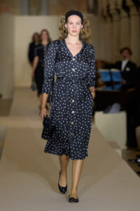 Model walking the runway in a navy polka-dot midi dress with a belt, headband, and matching flats at a fashion show.