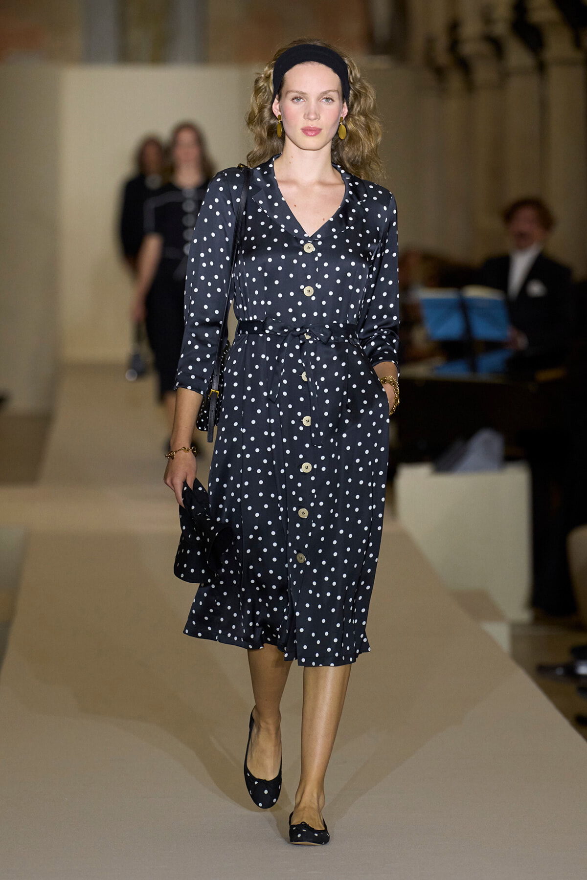 Model walking the runway in a navy polka-dot midi dress with a belt, headband, and matching flats at a fashion show.