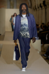 Male model walks a runway in a navy blue suit with a cloud-pattern shirt, carrying a navy-white tote bag