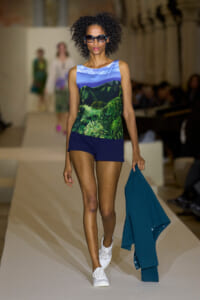 Model walking runway in a sleeveless landscape-print top, navy shorts, white sneakers, turquoise jacket in hand, oversized sunglasses.
