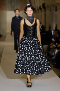Model on the runway wearing a black leather sleeveless bodice with buttons and a black and white polka-dot full skirt, plus a neck scarf and heels.