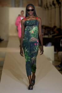 Model walking a fashion runway in a tropical-green printed jumpsuit with red straps, black sunglasses, and black platform sandals.