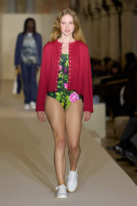 Model walking a runway in a red cardigan over a green floral one-piece, paired with white sneakers.
