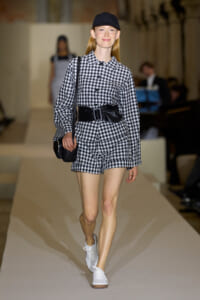 Female model walking a fashion runway in a black-and-white gingham romper with a wide black belt, cap, and white sneakers.