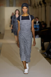 Runway model walking in a black‑and‑white gingham pinafore over a black top with white sneakers and a cap.
