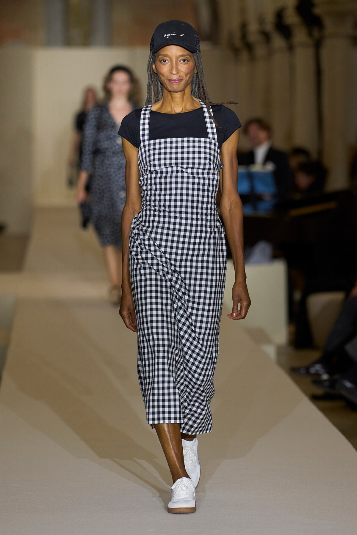 Runway model walking in a black‑and‑white gingham pinafore over a black top with white sneakers and a cap.