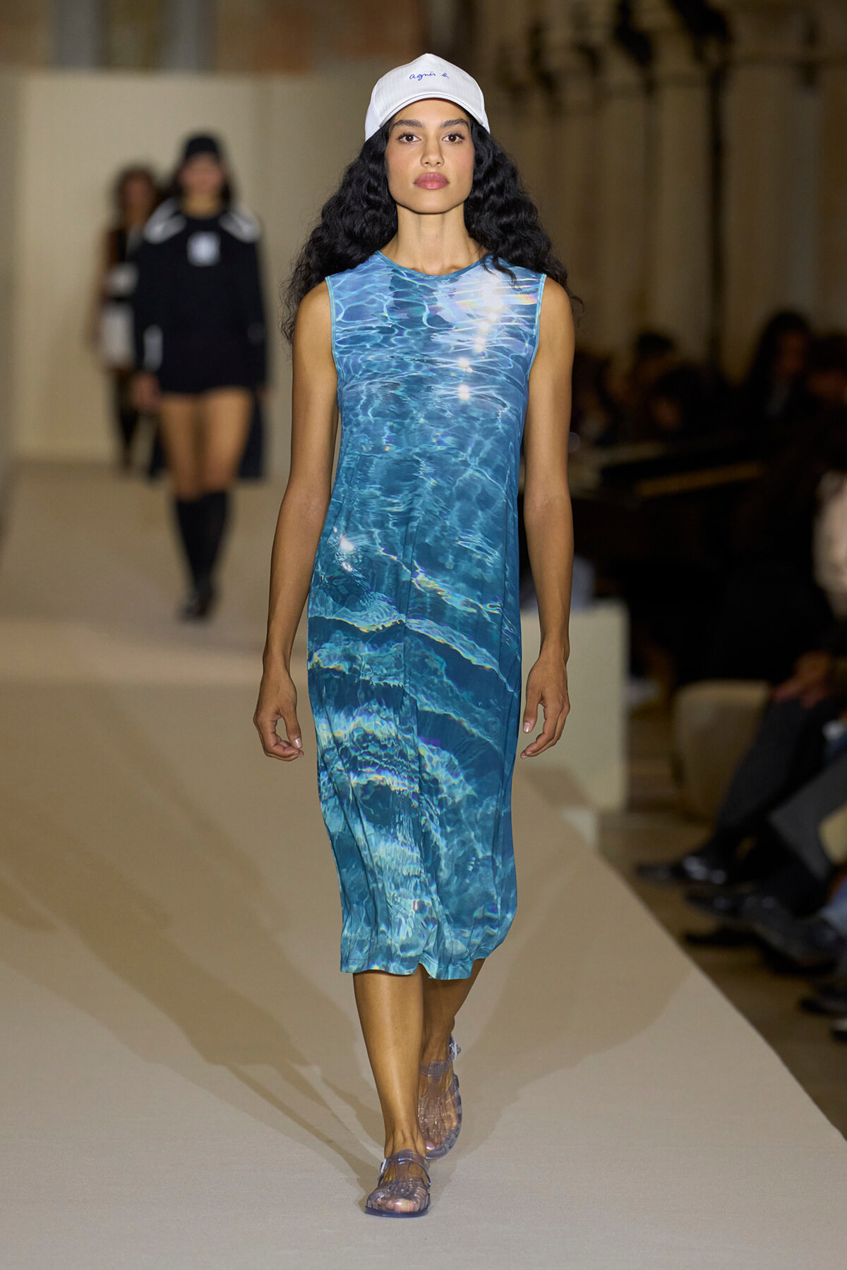 Model walks a runway wearing a sleeveless blue water-pattern midi dress and a white cap; she completes the look with clear sandals.