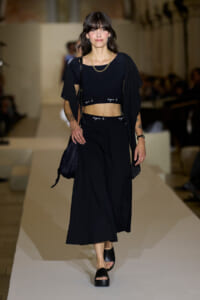 Model walking down a runway in all-black outfit: cropped top, wide-leg pants, cardigan, gold chain, and black sandals with a navy bag.