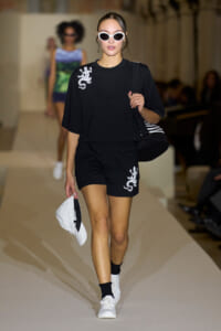 Model on a runway wearing a black top and shorts with white lizard embroidery, white sunglasses, a striped tote bag, and a white cap in hand.