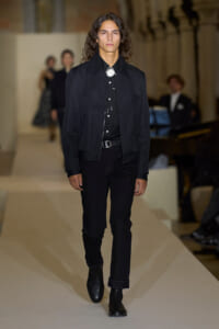 Male model walks the runway in an all-black outfit: black jacket, black shirt, black jeans, and black boots on a fashion show runway.